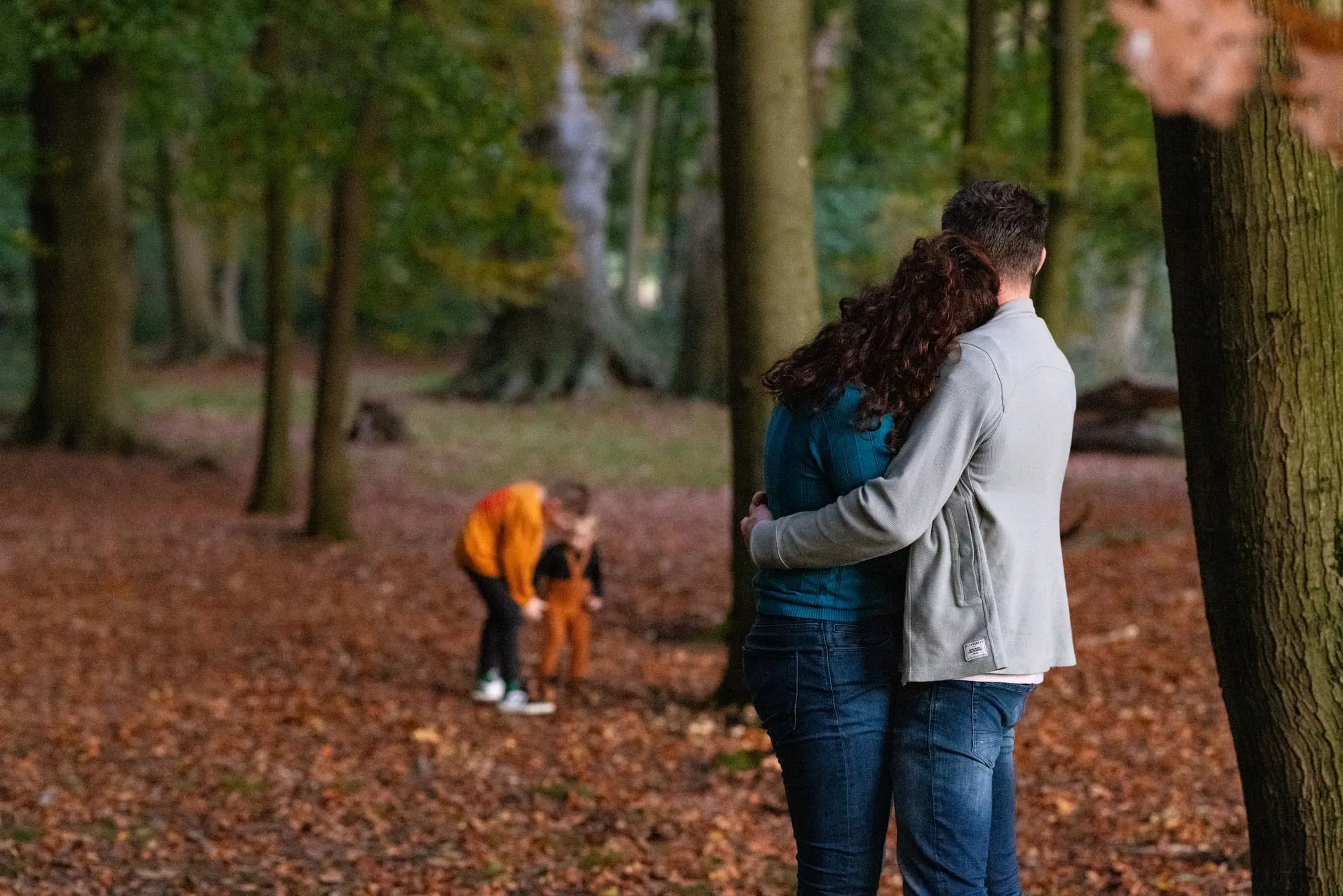 Ouders staan gearmd op een bospad terwijl hun kinderen verderop spelen