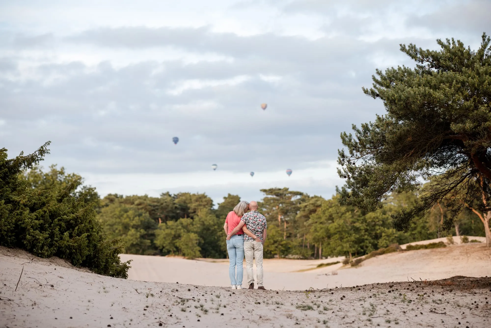Het stel staat klein in de zandverstuiving terwijl er luchtballonnen boven de bomen hangen