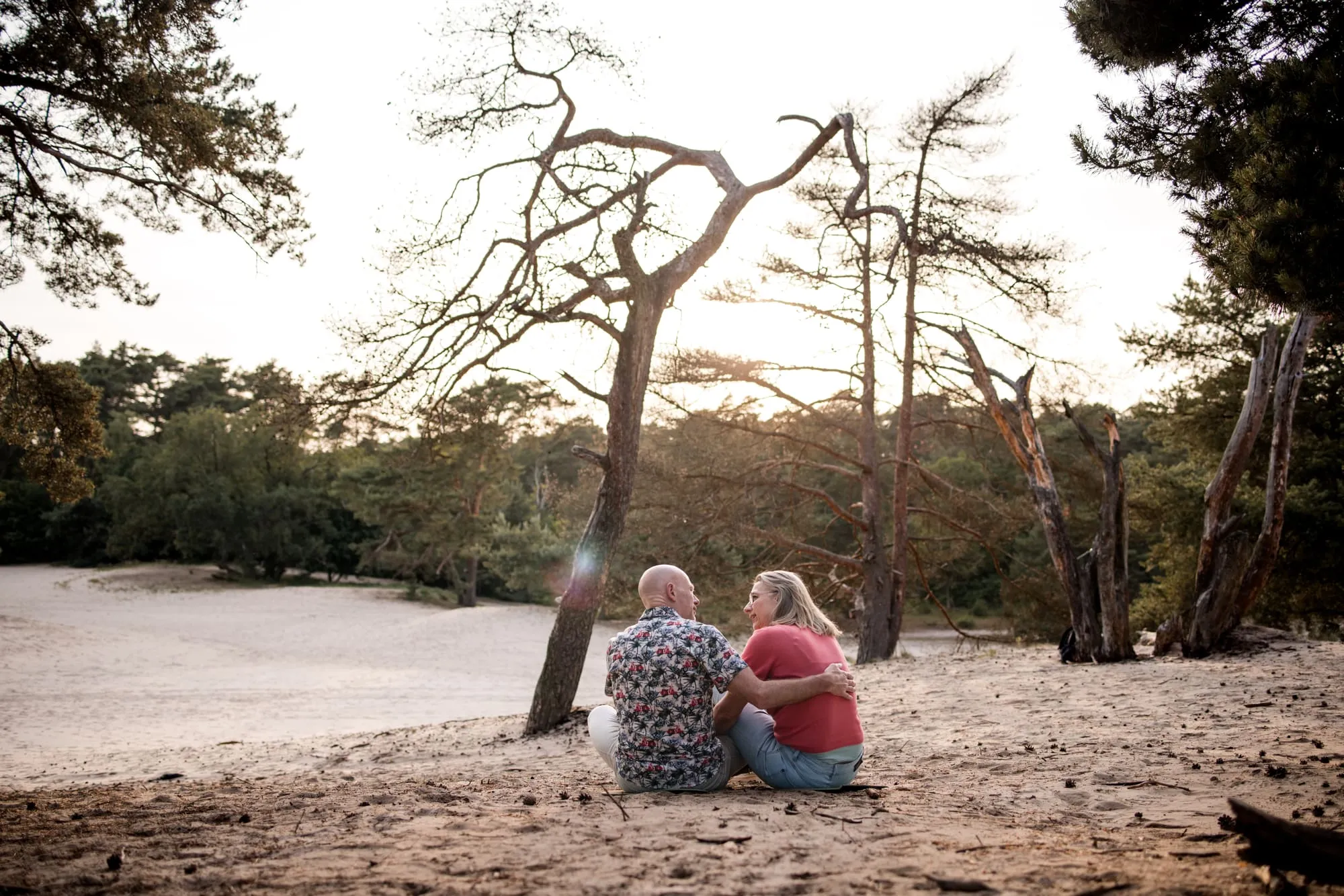 Het stel zit samen in het zand onder een oude den, met de zon laag achter de bomen