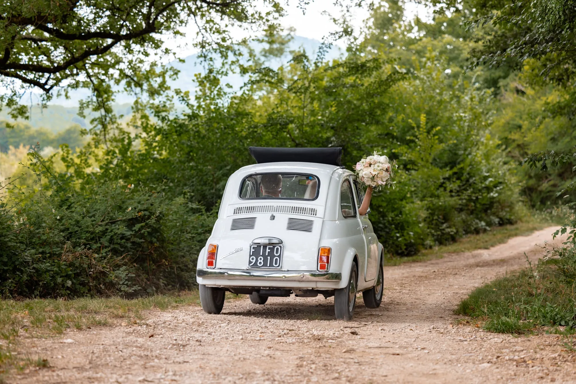 Een witte Fiat 500 rijdt weg over een onverhard pad, de bruid zwaait met haar boeket uit het raam