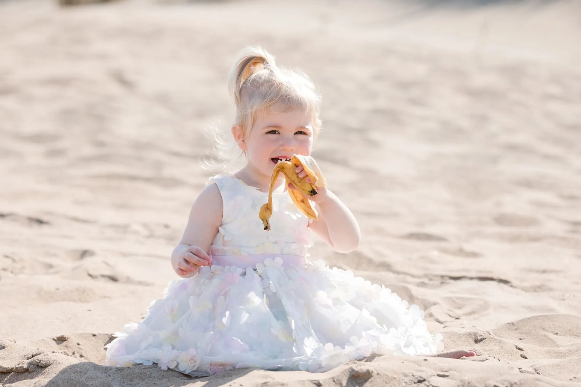 Een meisje in een tulen jurk eet een banaan in het zand